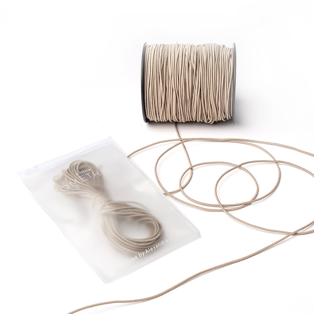 Eine beigefarbene Kordelspule mit loser Kordel und einem kleinen Elastikband-Bündel von Alexandra Renke in einem durchsichtigen Beutel, ideal für jedes Bastelprojekt, dargestellt auf weißem Hintergrund.