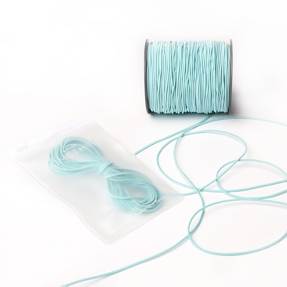 Eine Spule hellblaues Alexandra Renke Elastikband mit einem passenden Bündel in einem durchsichtigen Beutel, perfekt für Ihr nächstes Bastelprojekt, auf weißem Hintergrund.