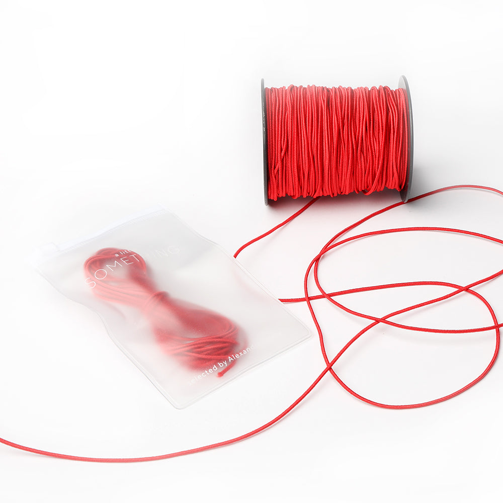 Alexandra Renke rote Kordel auf einer Spule, dazu lose Kordel und ein kleineres Bündel in einem transparenten Beutel - das perfekte Elastikband für jedes Bastelprojekt - auf weißem Hintergrund.