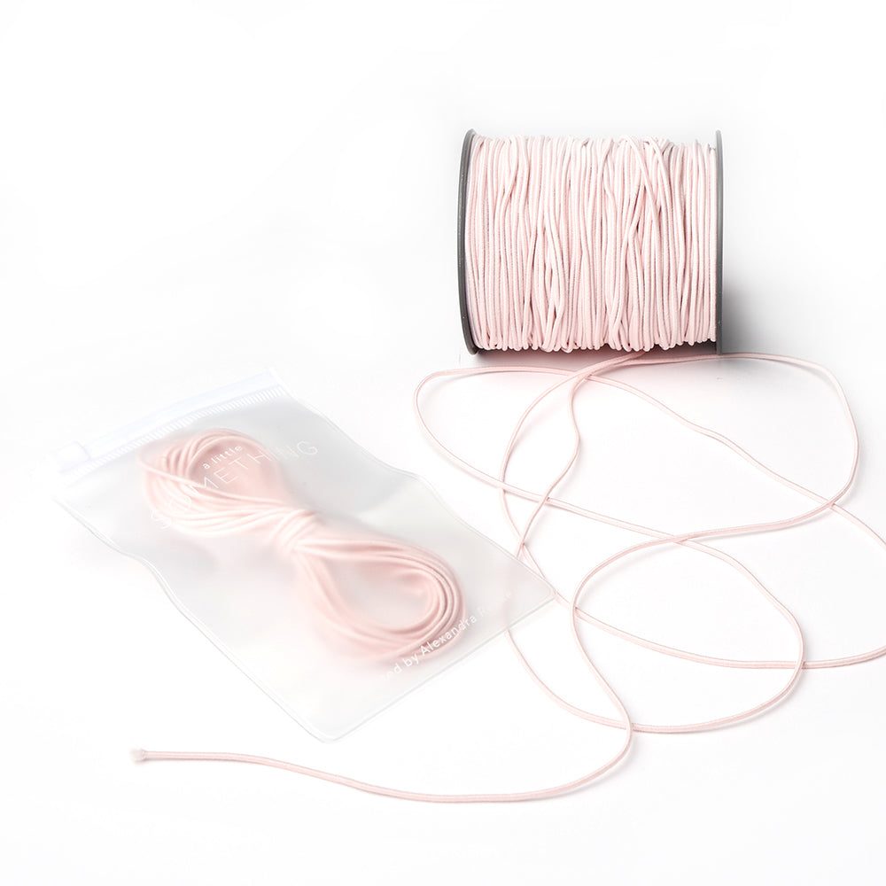 Eine Spule hellrosa Elastikband von Alexandra Renke, mit etwas abgewickelter Kordel und einem versiegelten Beutel mit zusätzlicher rosa Kordel - ideal für jedes Bastelprojekt.