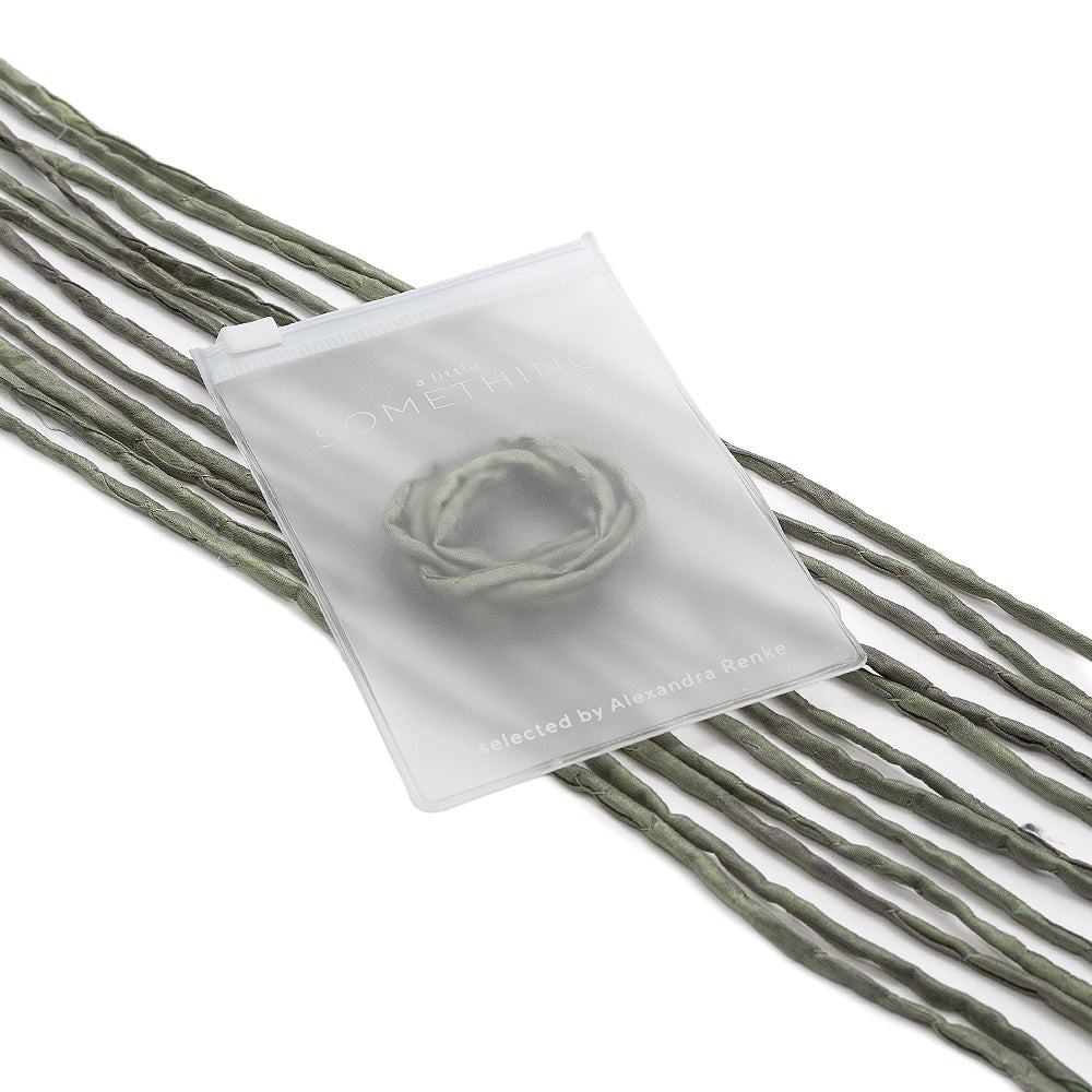 Ein mattes Säckchen mit einem gewundenen Gegenstand darin, das auf mehreren langen, grünen, gedrehten Seidenschnur-Kordeln von ☆ präsentiert wird – ideal für einzigartige Geschenkverpackungen oder als bezauberndes, handgenähtes Detail.