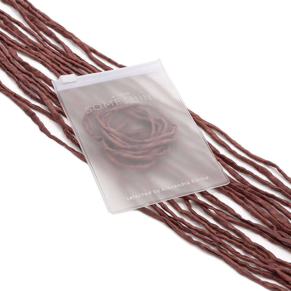 Ein durchscheinender Beutel mit Schriftzug, handgenäht von ☆, liegt auf braunen Garnsträngen, die in einem wellenförmigen Muster angeordnet und durch eine zarte Seidenschnur akzentuiert sind – perfekt für stilvolle Geschenkverpackungen.