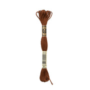 Ein Knäuel braunes DMC Mouliné Garn, etikettiert mit 801 und umwickelt mit creme- und goldfarbenen Bändern. Hergestellt aus 100% Baumwolle, ist dieses Stickgarn perfekt für Stickereiprojekte.