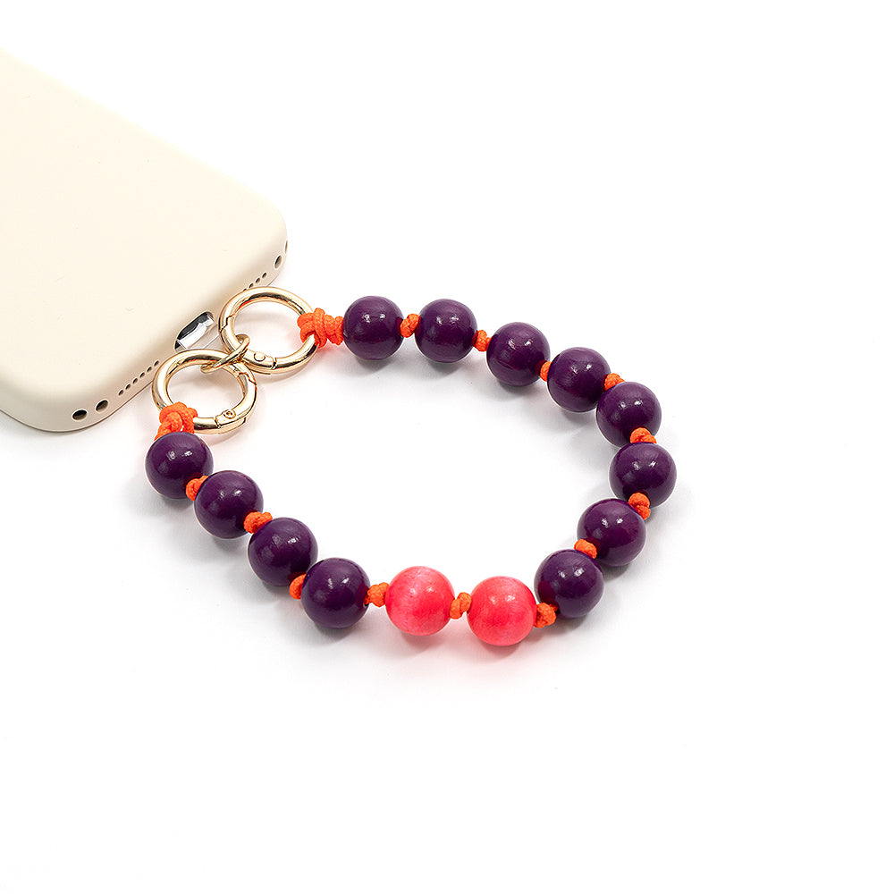 Eine cremefarbene Handyhülle von ☆ mit einer Handschlaufe aus violetten und rosa Holzperlen an einer orangefarbenen Flechtkordel.