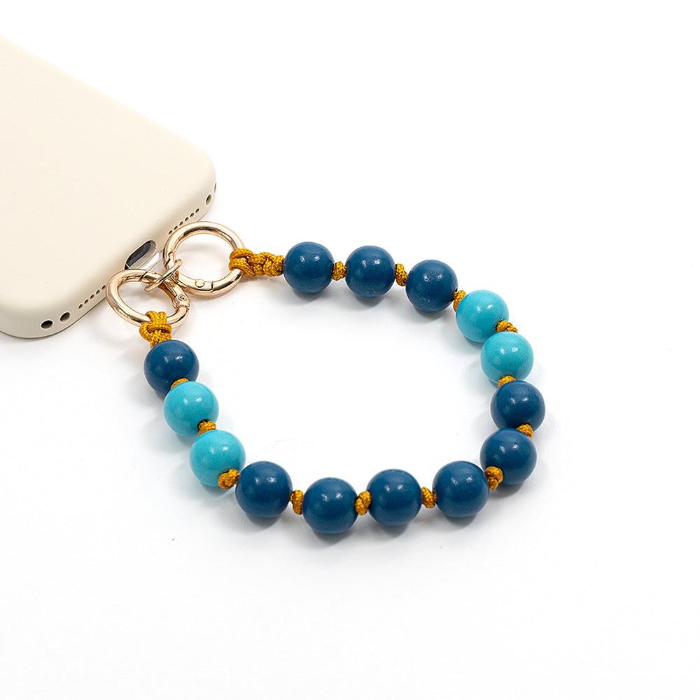 Ein weißes Smartphone ist an einem ☆ blau-türkisfarbenen Makramee-Armband aus Holzperlen mit goldenen Ringverschlüssen befestigt und wird vor einem weißen Hintergrund präsentiert.