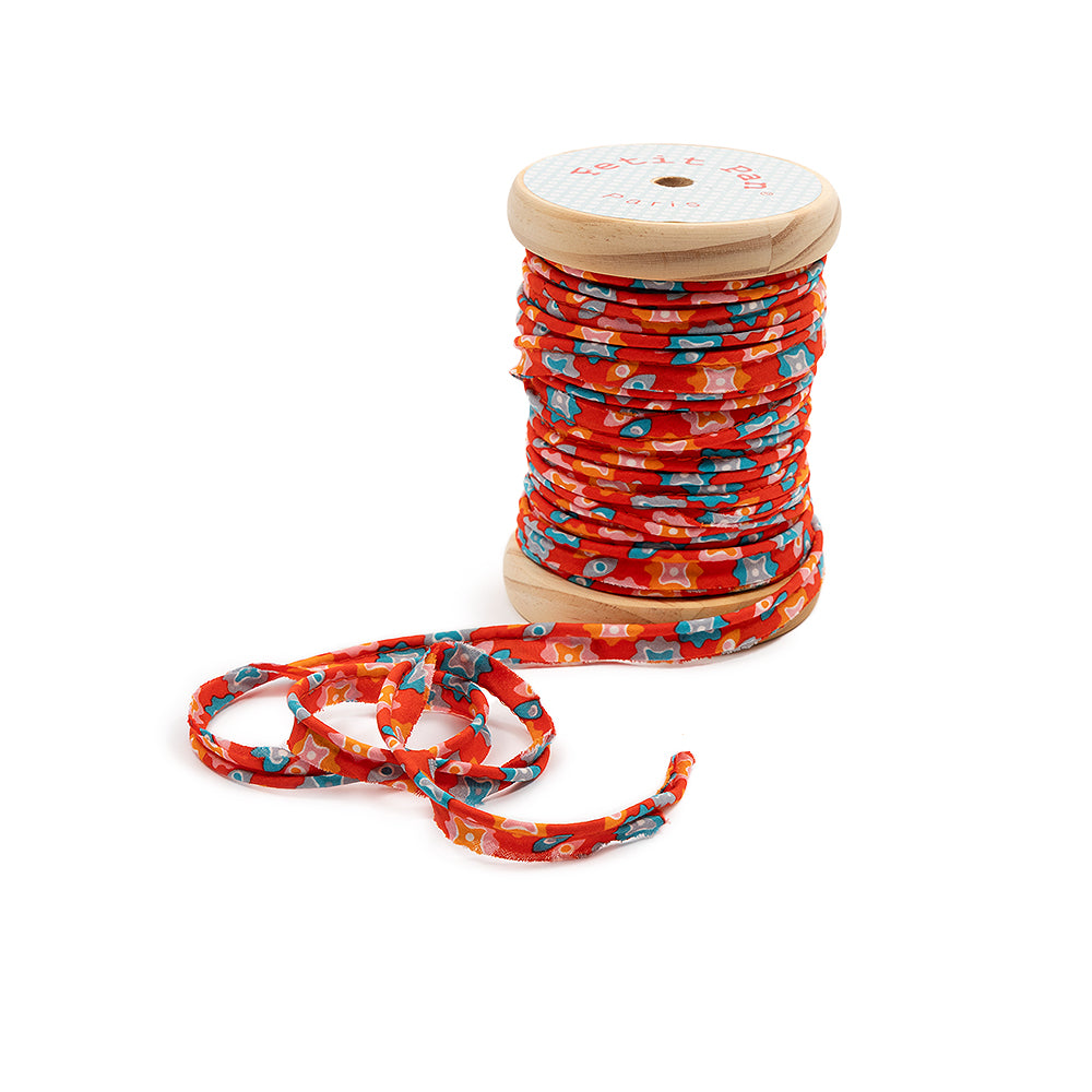 Eine Holzspule mit rot, blau, orange und weiß gemustertem Schrägband – ideal zum Nähen – teilweise abgewickelt und vorne aufgerollt, perfekt, um Stoffkanten mit einer dekorativen Note zu veredeln.