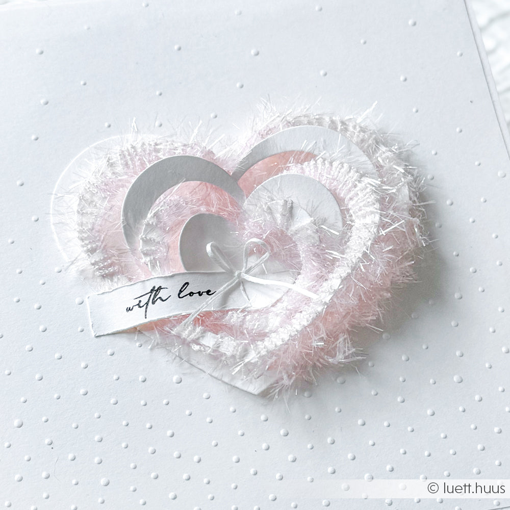Handgefertigte Karte mit übereinander liegenden rosa und weißen Herzen, einem Liebesanhänger und strukturierten Punkten - hergestellt mit Rico Design Creative Bubble Garn für einen einzigartigen Touch auf weißem Hintergrund.