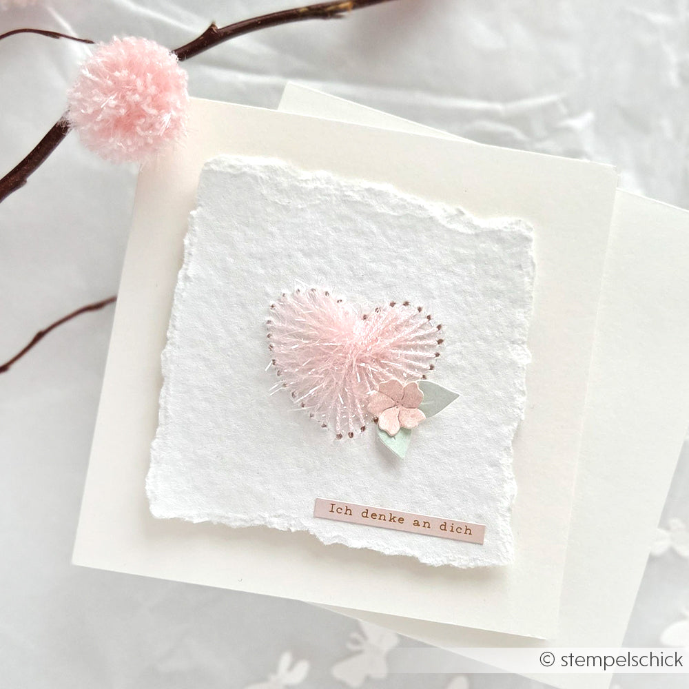 Handgemachte Karte von Rico Design mit einem rosa Herz aus robustem Garn, kleinen Papierblumen und dem deutschen Text "Ich denke an dich.