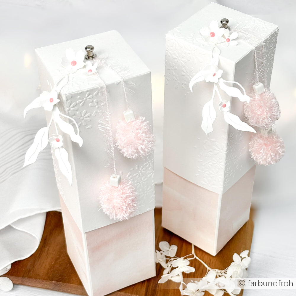 Zwei elegante Geschenkschachteln von Rico Design in Weiß und Rosa, verziert mit flauschigen Bommeln, Papierblumen, geprägten Mustern und veredelt mit robustem Garn für einen einzigartig charmanten Look.