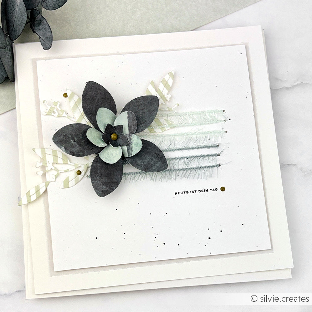 Quadratische handgefertigte Karte von Rico Design mit einer geschichteten schwarzen und weißen Papierblume, strukturierten Akzenten und Creative Bubble Garn-Details auf einem strahlend weißen Hintergrund.