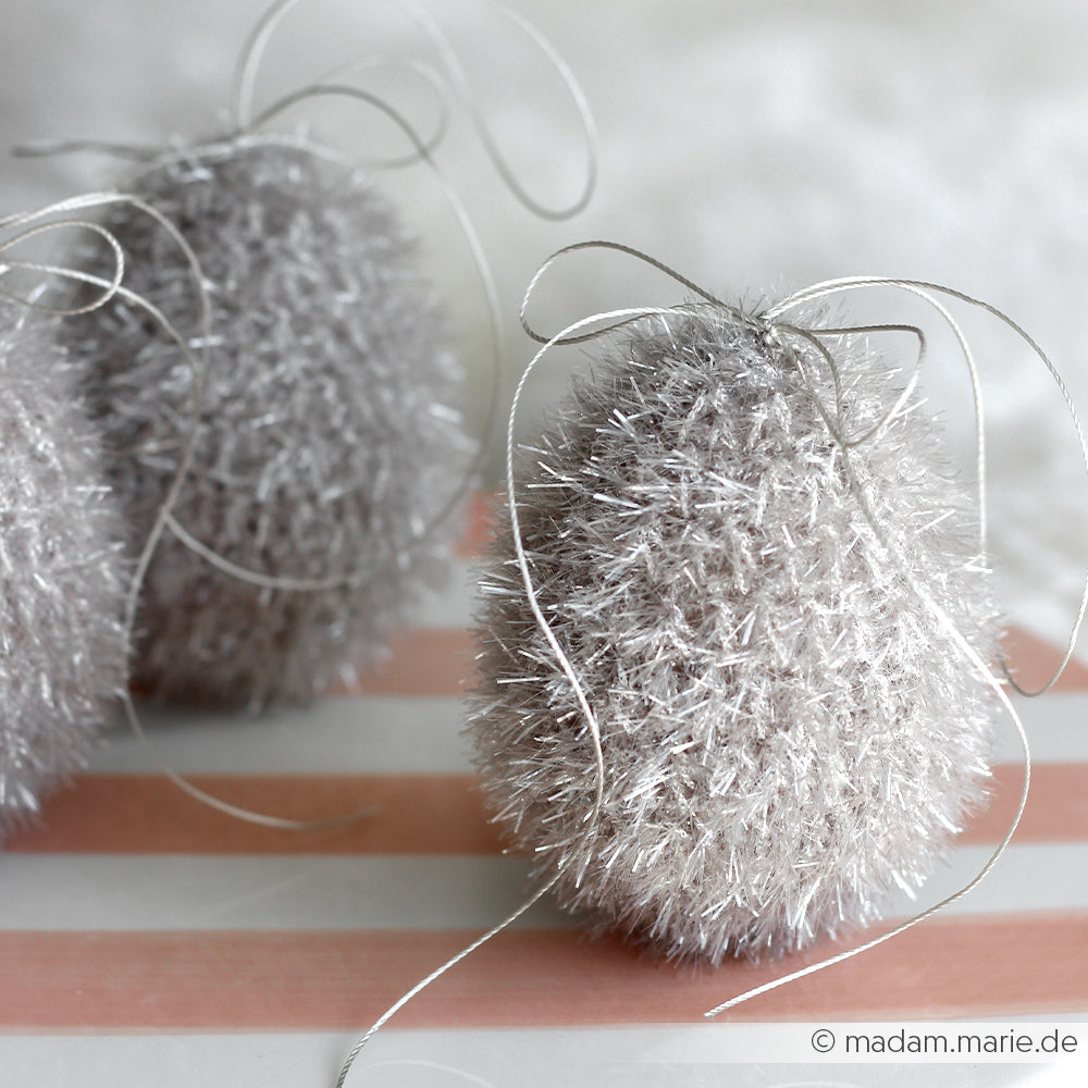 Drei flauschige, silberne, eiförmige Dekorationen, gehäkelt mit Rico Design Creative Bubble Garn und mit Bändern verziert, ruhen auf einer rosa-weiß gestreiften Fläche.