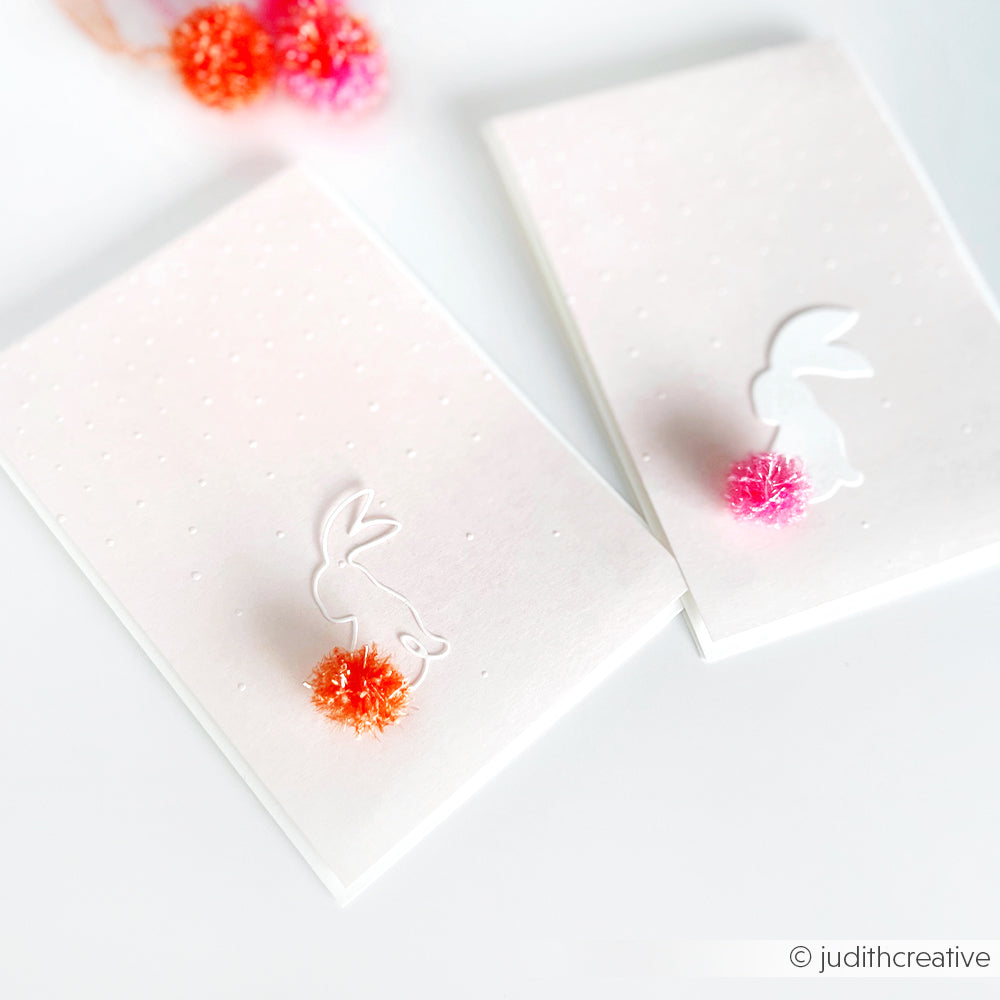 Zwei weiße Karten mit Häschensilhouetten und bunten Bommelschwänzen - eine orange, eine rosa - aus Rico Design Creative Bubble Garn, die auf einer weißen Fläche präsentiert werden. Ideal für verspielte Dekoration oder Häkeln Spüllappen Projekte.