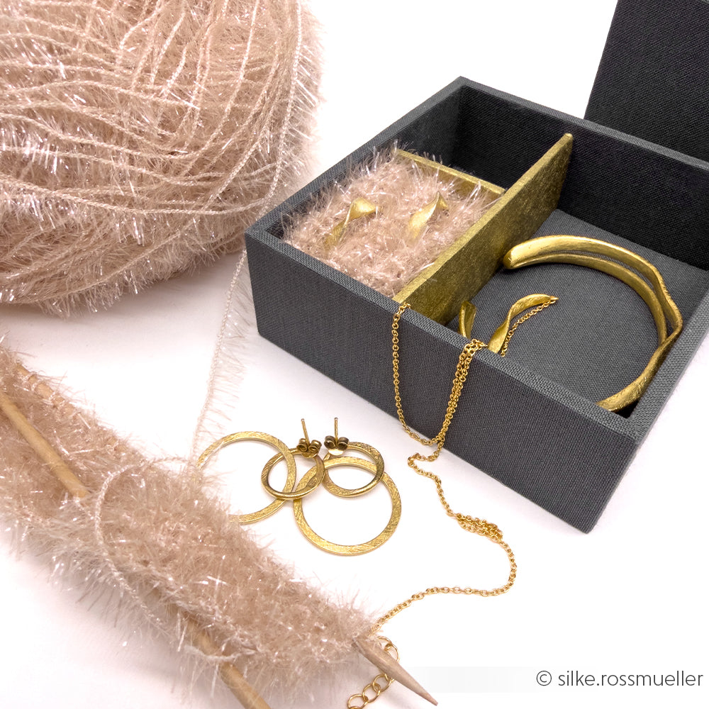 Goldschmuck von Rico Design in einer grauen Schachtel mit flauschigem beigem Garn, robustem Garn zum Häkeln von Geschirrtüchern, Stricknadeln und drei Paar Ohrringen daneben.