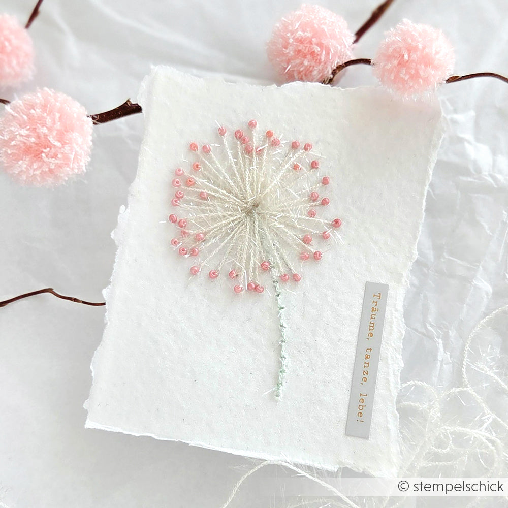 Handgefertigte Karte von Rico Design mit einer gestickten Pusteblume mit robustem Garn und rosafarbenen Perlen, umgeben von rosafarbenen Bommelzweigen - eine ideale Inspiration für kreative Projekte wie das Häkeln von Geschirrtüchern.