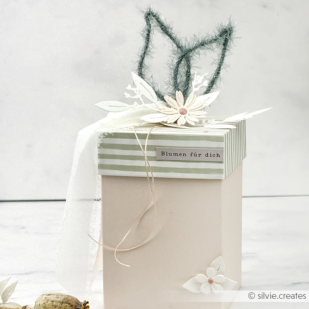 Eine pastellfarbene Geschenkschachtel von Rico Design mit Blumendekoration, Hasenohren, einem "Blumen für dich"-Etikett und robustem Garn für eine charmante Note.