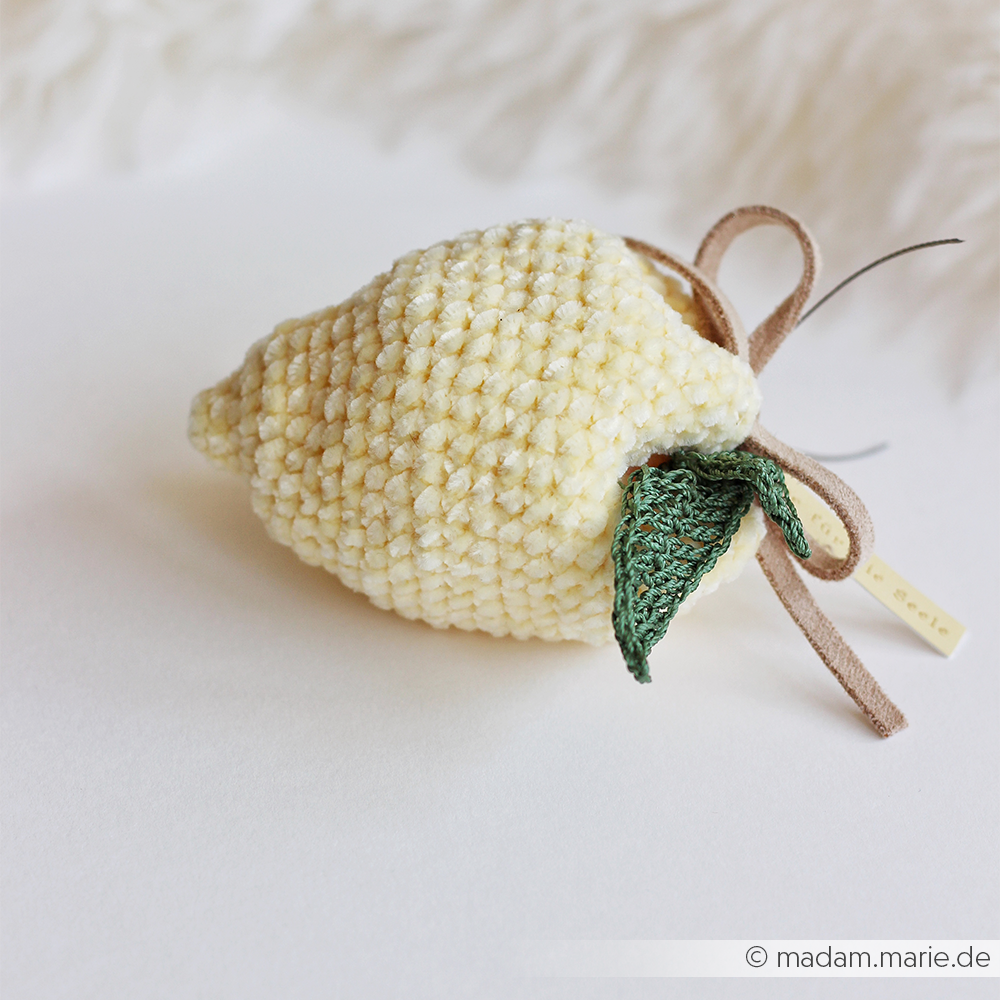 Eine gehäkelte Zitrone mit grünen Blättern und einem braunen Band von Rico Design, hergestellt aus superflauschigem Chenille-Garn, ausgestellt auf einer weißen Fläche.