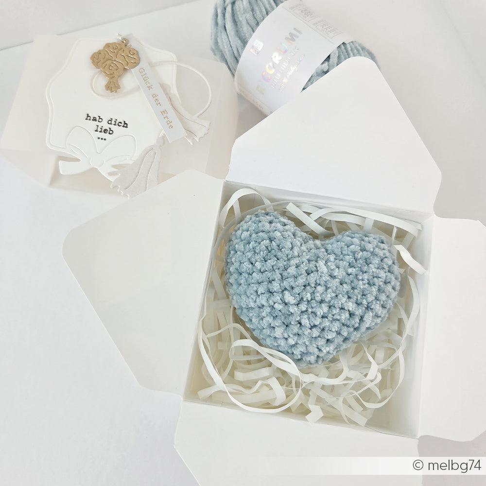 Ein blaues gehäkeltes Herz aus weichem Chenille Garn von Rico Design ist in einer weißen Geschenkbox mit geschreddertem Papier und einem Garnetikett untergebracht, ideal für Bastelprojekte oder Häkeln für Babys.