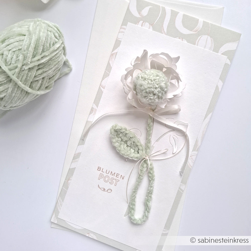 Eine handgefertigte Karte mit einer Blume und Blättern aus strukturiertem Garn, die neben einem Knäuel weichem grünen Chenillegarn von Rico Design platziert ist.