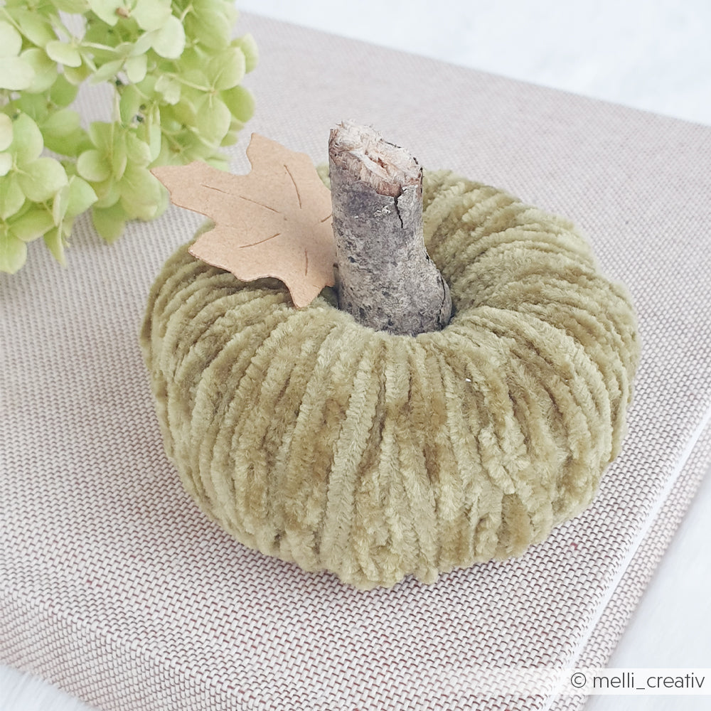 Ein weicher, grüner Samtkürbis mit einem Zweig als Stiel und einem Blatt aus Papier steht auf einer strukturierten, beigen Stoffoberfläche, die an kuschelweiche Tücher aus Chenille-Garn erinnert.