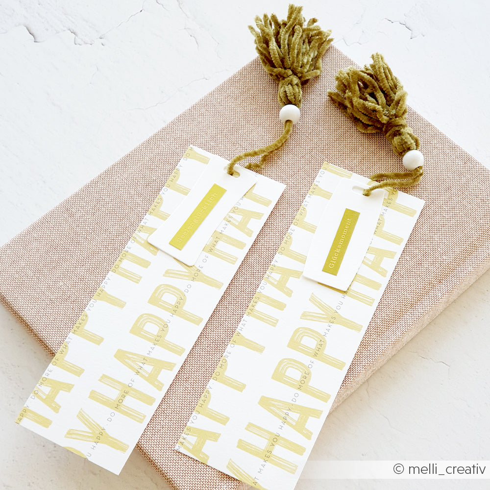 Zwei HAPPY-Lesezeichen von Rico Design mit Quasten und Perlen aus superflauschigem Garn liegen auf einem strukturierten beigen Bucheinband.