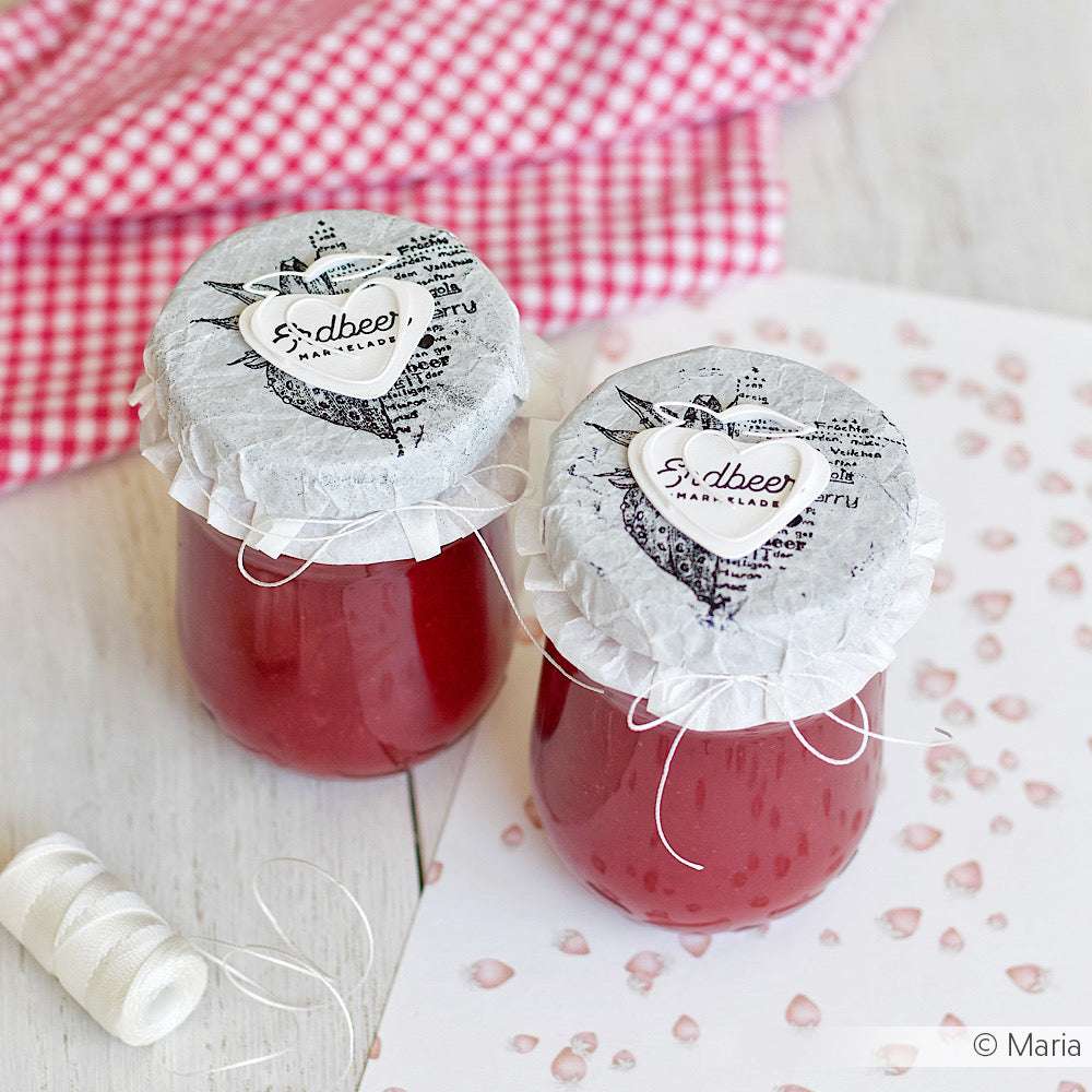 Zwei Gläser mit roter Marmelade von Alexandra Renke, die mit Papierdeckeln und Schnüren versehen sind, die an Kumihimo oder Makramee-Flechtung erinnern, werden auf einem Hintergrund aus Gingham-Stoff präsentiert.