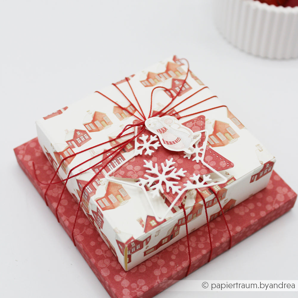 Zwei übereinander gestapelte Weihnachtsgeschenke, eingepackt in Papier mit Hausmotiv und mit rotem Kumihimo-Band verschnürt.