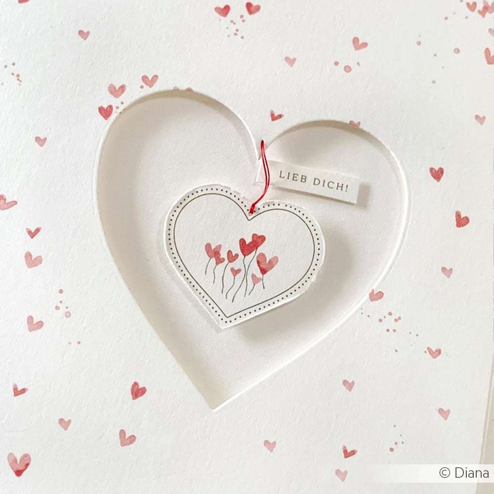 Weiße Karte von Alexandra Renke mit einem Herzausschnitt, roten Herzen im Inneren und einem "Lieb Dich!"-Anhänger - perfekt in Kombination mit einer Perlenschnur oder einem Makramee-Armband für ein herzliches Geschenk.