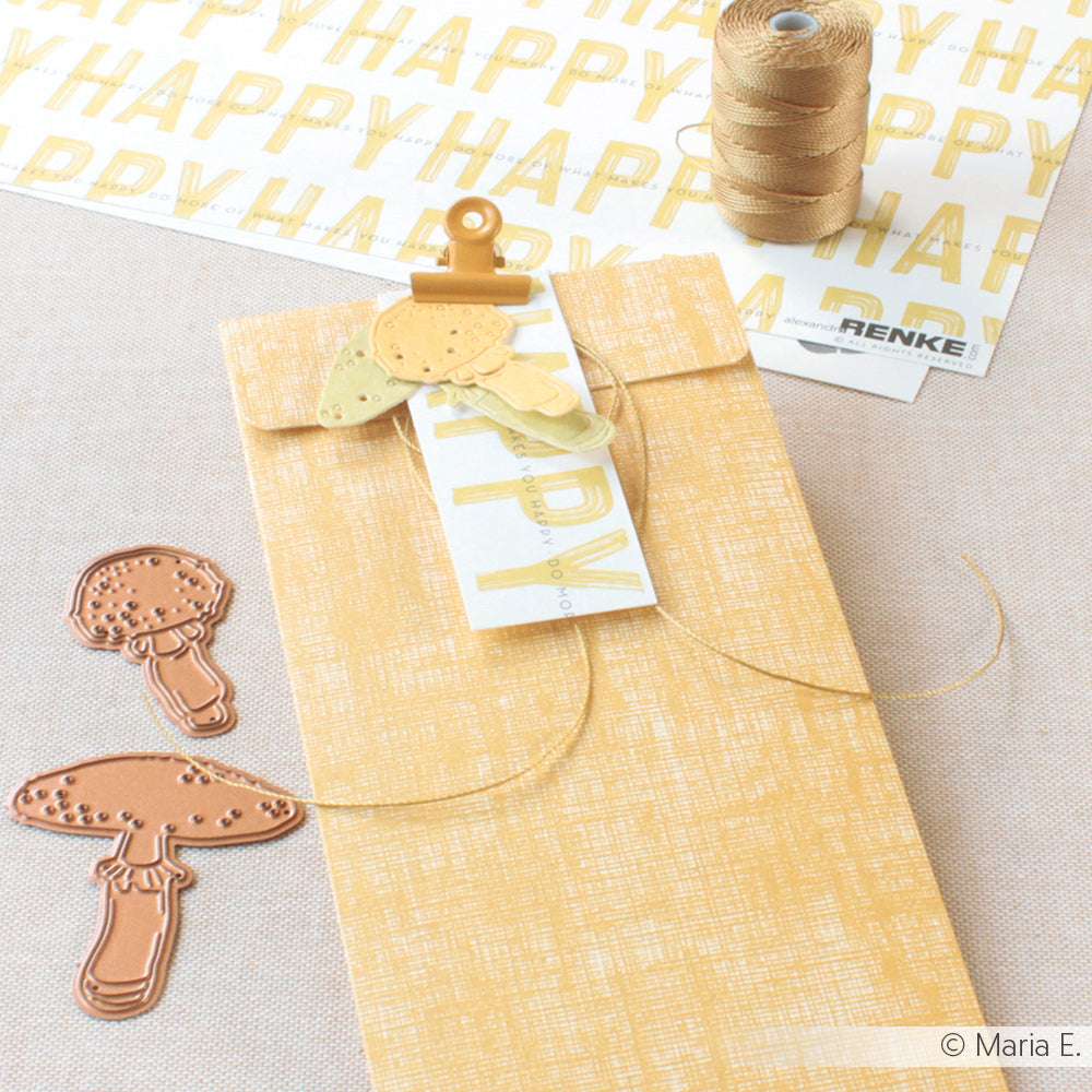 Ein gelber Geschenkumschlag von Alexandra Renke mit Schnur, Pilzausschnitten, Goldfaden und einem mit zarter Perlenschnur verzierten HAPPY-Anhänger liegt auf einer hellen Fläche.