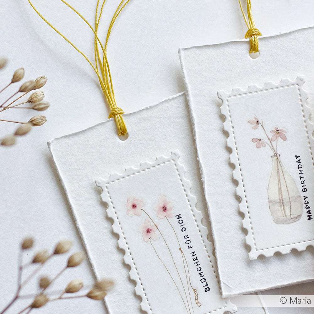 Zwei handgefertigte Geschenkanhänger von Alexandra Renke mit zarten floralen Illustrationen und gelben Kumihimo-Schnüren, die auf einer weißen Fläche präsentiert werden.