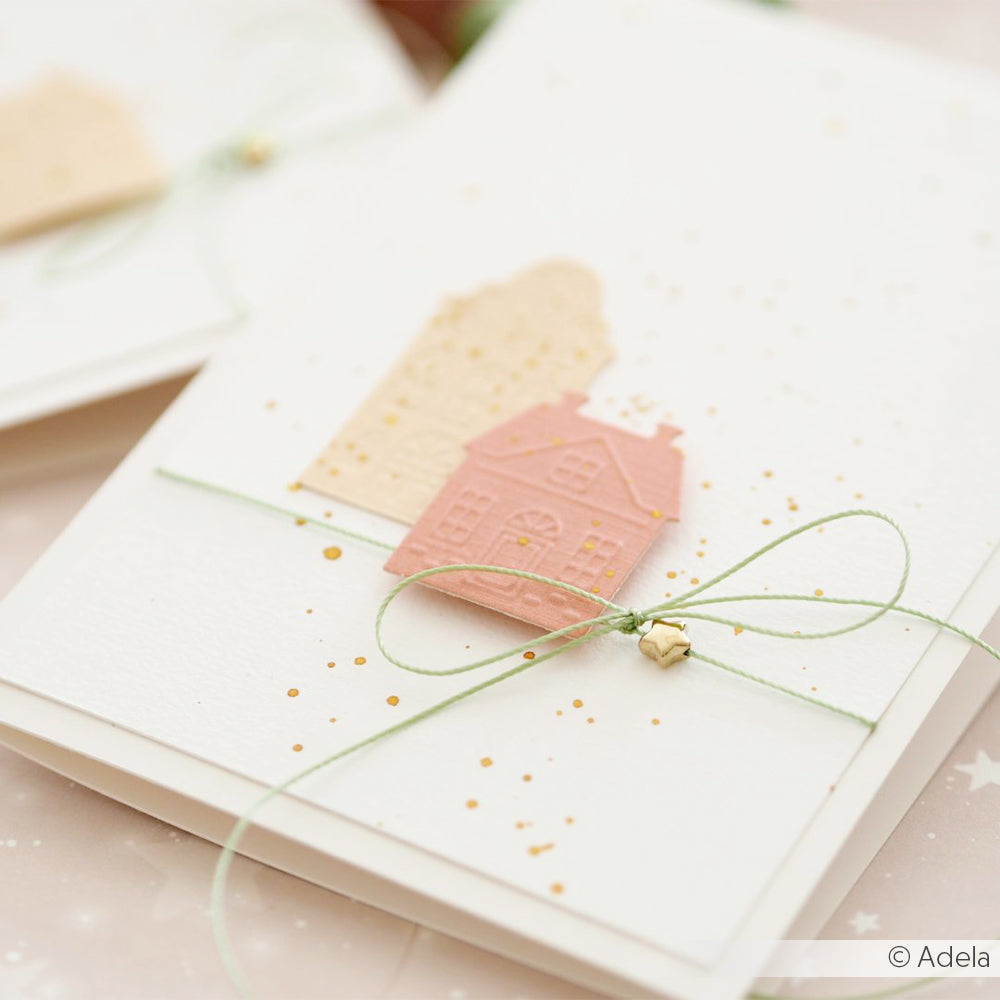 Handgefertigte Grußkarte von Alexandra Renke mit pastellfarbenen Papierhäusern, einer grünen Schleife im Makramee-Stil und goldenen Sprenkeln auf weißem Hintergrund.
