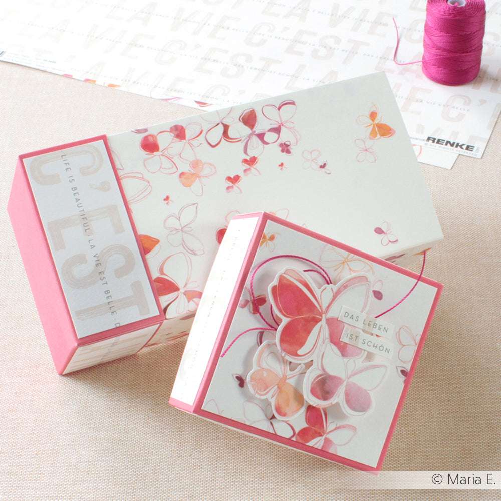 Zwei floral gemusterte Geschenkboxen von Alexandra Renke mit Schmetterlingsmotiven und deutschem Text, die neben violettem Garn ausgestellt sind - ideal, um Kumihimo- oder Makramee-Kreationen zu präsentieren.