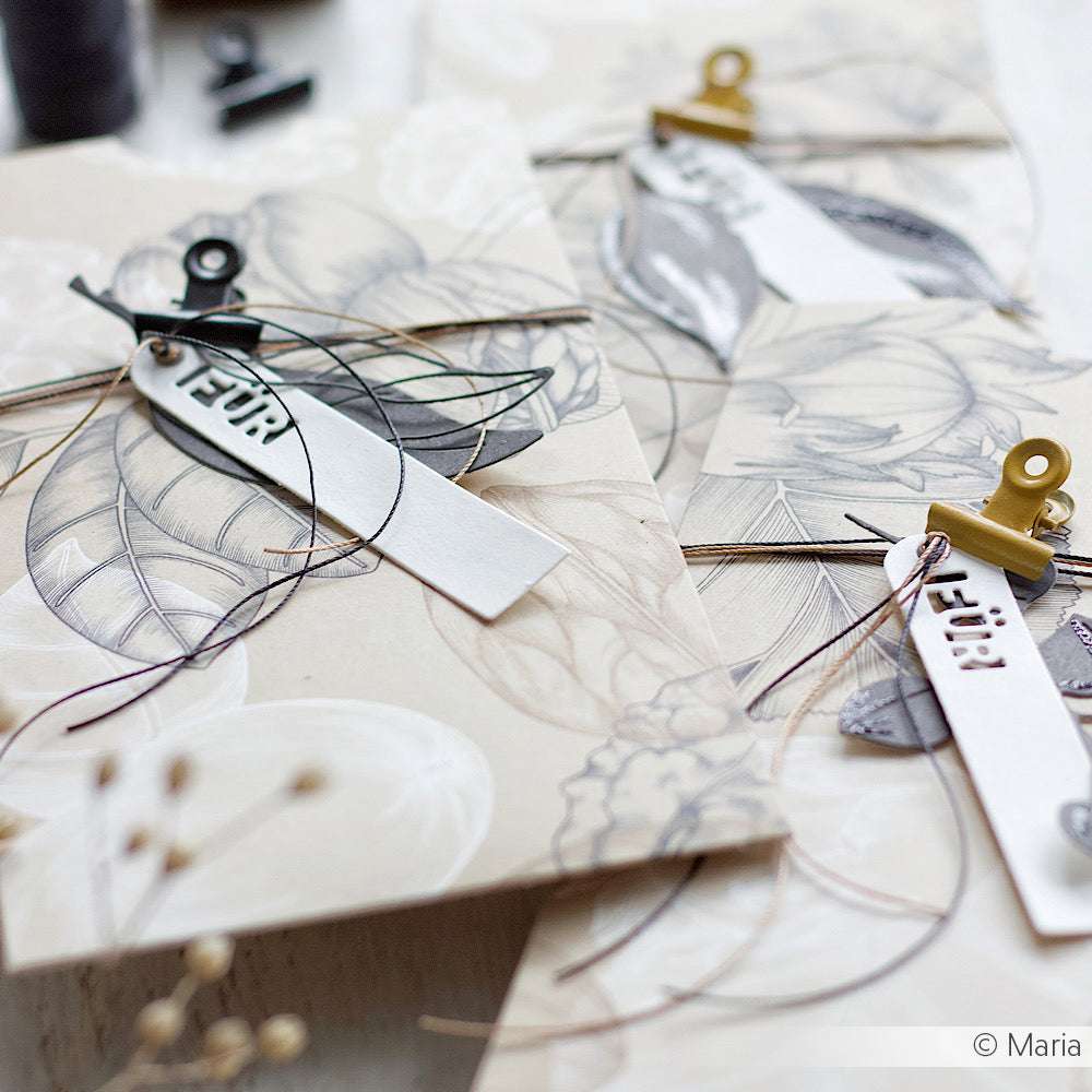 Die von Alexandra Renke handgefertigten Karten mit floralen Motiven, Makramee-Schnur und weißen Anhängern, die von Klammern gehalten werden, sind kunstvoll auf einer hellen Fläche arrangiert.