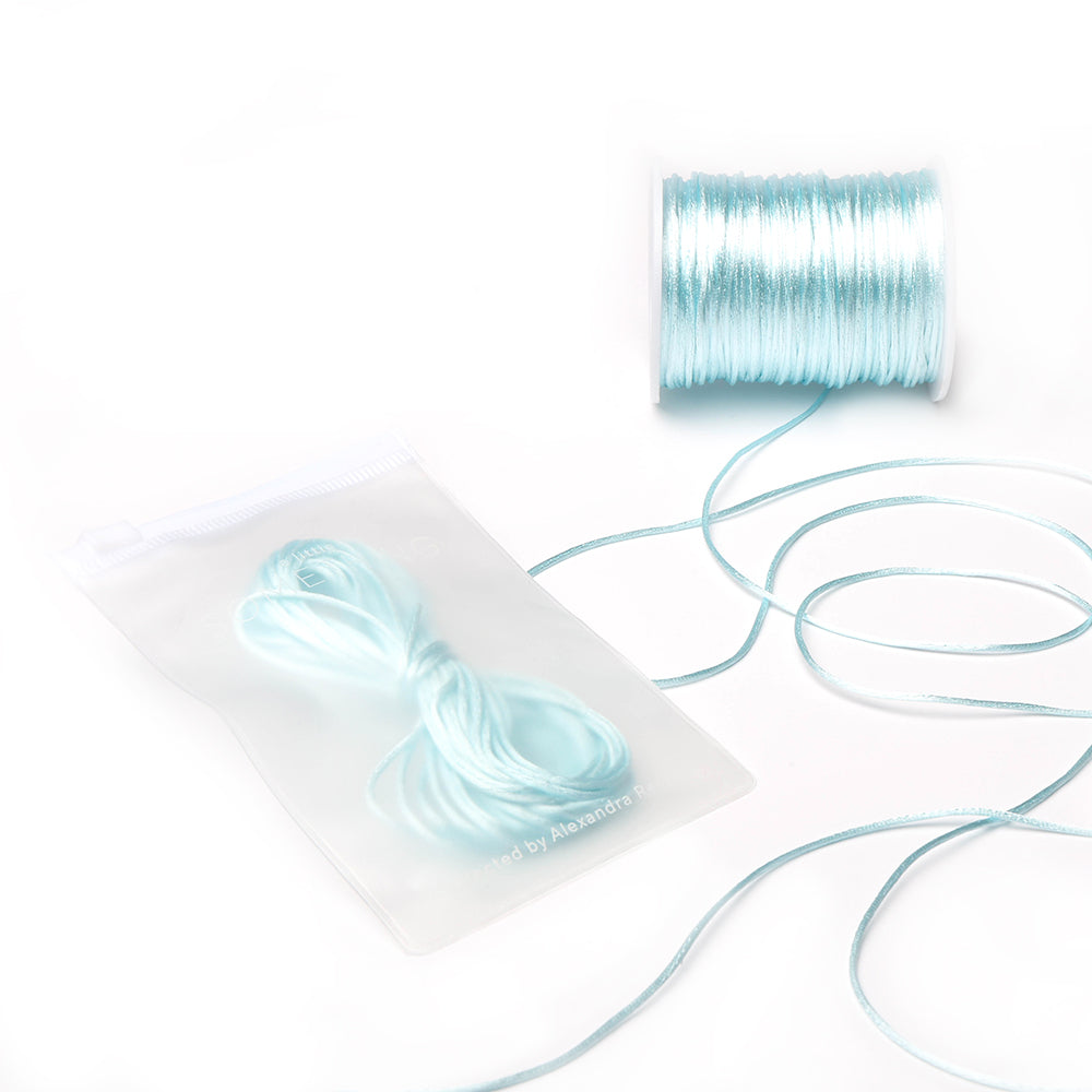 Eine Spule und ein Bündel hellblauer Satinkordeln von Alexandra Renke in einem durchsichtigen Reißverschlussbeutel auf weißem Hintergrund – ideal für DIY-Wohnaccessoires oder zum Herstellen von Schmuck.