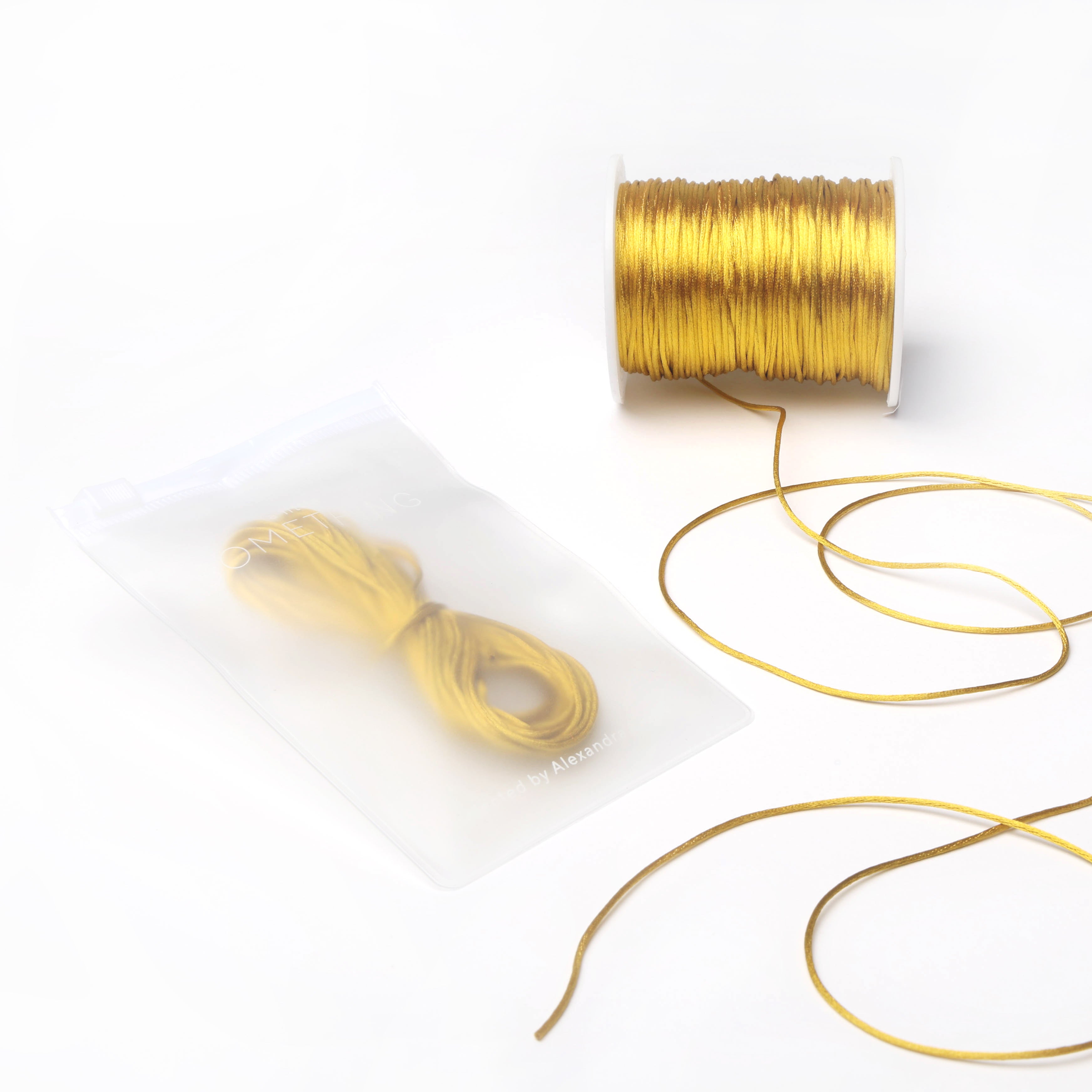 Eine Spule mit goldenen Satinkordeln von Alexandra Renke, daneben eine lose Kordel und eine Packung mit weiteren goldenen Kordeln – perfekt für DIY-Projekte zur Wohnraumgestaltung oder zur Schmuckherstellung.