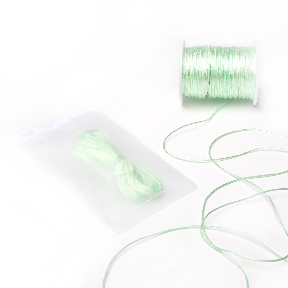 Eine Spule und ein Bündel hellgrüner Satinkordeln von Alexandra Renke, wobei ein Bündel in einer halbtransparenten Plastiktüte verpackt ist – ideal für DIY-Projekte im Bereich Wohnkultur oder zur Herstellung von Schmuck.