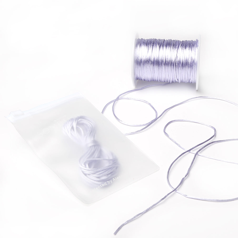 Neben einem durchsichtigen Beutel, in dem sich ein aufgerolltes Stück derselben Kordel befindet, liegt eine Spule mit helllavendelfarbenen Satinkordeln von Alexandra Renke – ideal für DIY-Wohnaccessoires oder alle, die Schmuck mit einem eleganten Satin-Touch herstellen möchten.