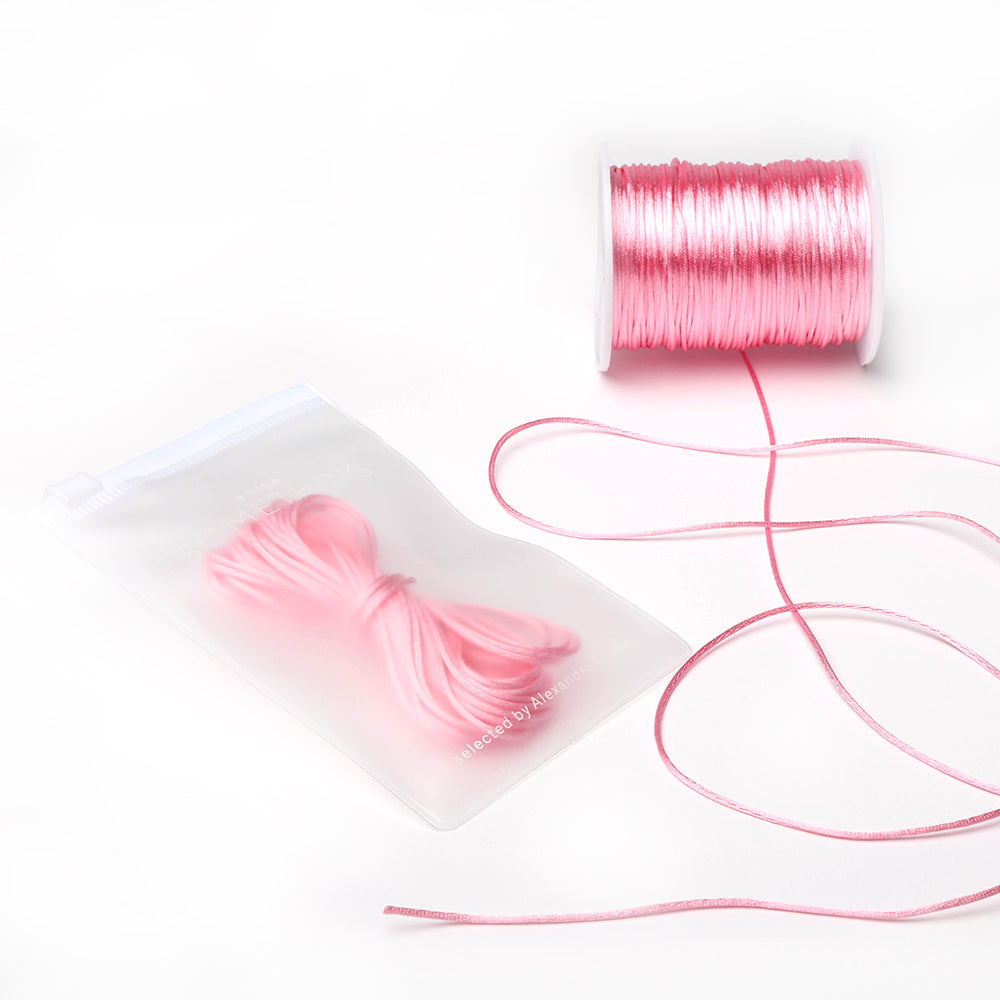 Eine Spule und ein Bündel glänzender rosa Satinkordeln von Alexandra Renke, ideal zum Schmuck basteln oder für DIY-Wohnaccessoires, wobei das Bündel ordentlich in einem durchsichtigen Plastikbeutel mit Reißverschluss verpackt ist.