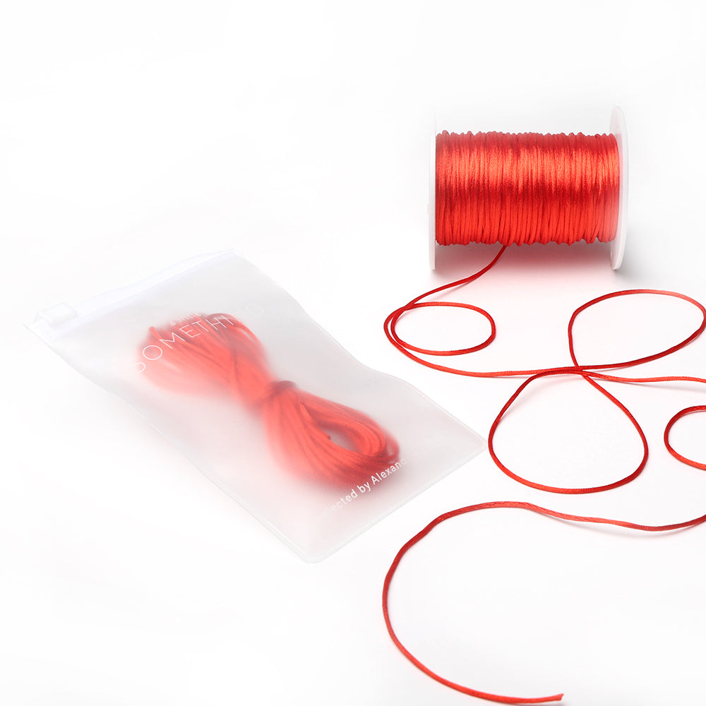 Alexandra Renke: Rote Satinkordeln auf einer Spule, dargestellt als aufgerolltes Bündel in einem halbtransparenten Reißverschlussbeutel – ideal für DIY-Wohnaccessoires oder die Schmuckherstellung –, präsentiert vor weißem Hintergrund.