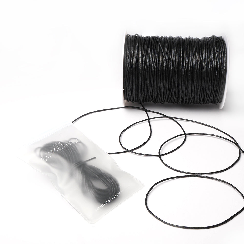 Eine Spule schwarzer gewachster Baumwollkordel von Alexandra Renke, mit einem Abschnitt in einer durchsichtigen Plastiktüte, ideal für Schmuckherstellung oder Dekoration, ausgestellt auf weißem Hintergrund.