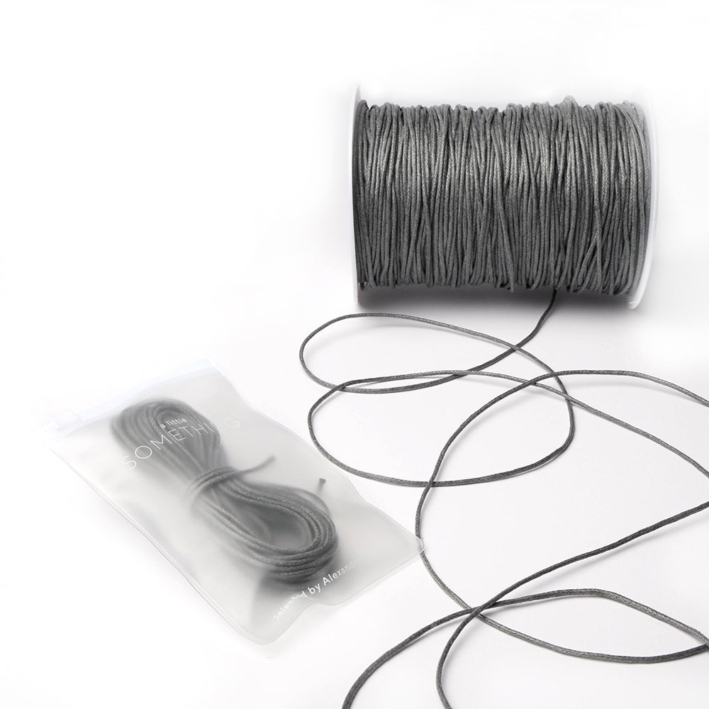 Eine Spule grau gewachste Baumwollkordel von Alexandra Renke, etwas abgewickelte Kordel und ein kleineres Bündel in einem durchsichtigen Beutel - ideal für Schmuckherstellung oder Dekorationsprojekte.