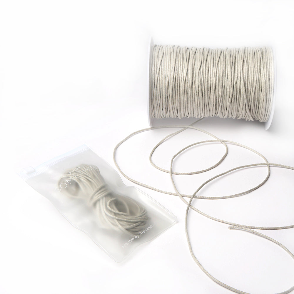 Eine Spule und ein kleines Bündel beigefarbener, gewachster Baumwollkordel von Alexandra Renke mit losen Strängen auf weißem Hintergrund, ideal für Dekoration oder Schmuckherstellung.