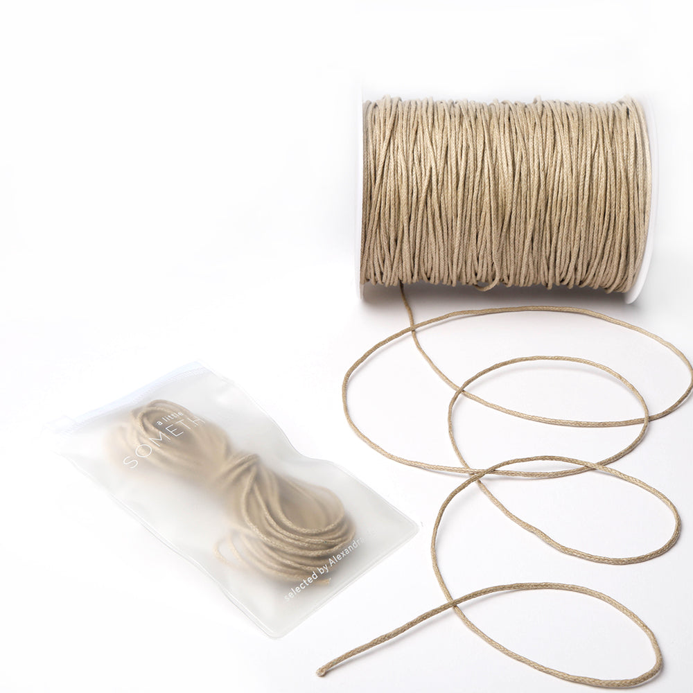 Eine Spule beige gewachste Baumwollkordel von Alexandra Renke mit einer losen Länge, daneben ein versiegelter Beutel mit mehr - ideal für Dekorations- oder Schmuckherstellungsprojekte.