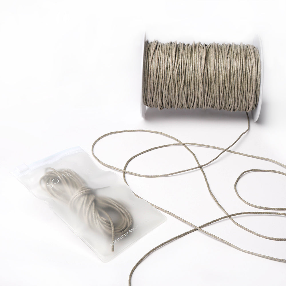 Eine Spule der beigefarbenen, gewachsten Baumwollkordel von Alexandra Renke, teilweise abgewickelt, liegt neben einem kleineren Bündel in einem halbtransparenten Beutel - ideal für Dekorations- oder Schmuckherstellungsprojekte.