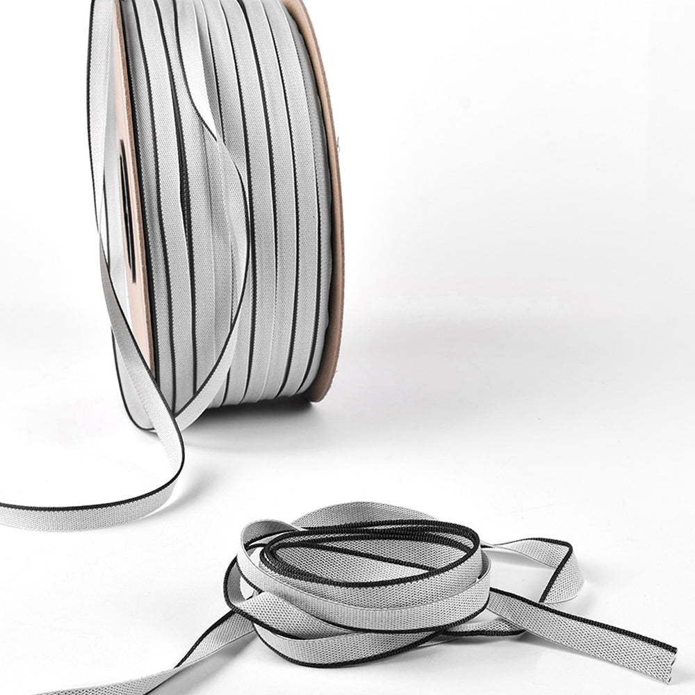 Eine Spule mit grau-schwarzem, reflektierendem Klebeband von Alexandra Renke, daneben lose aufgerollte Bänder, ruht auf einem weißen Hintergrund.