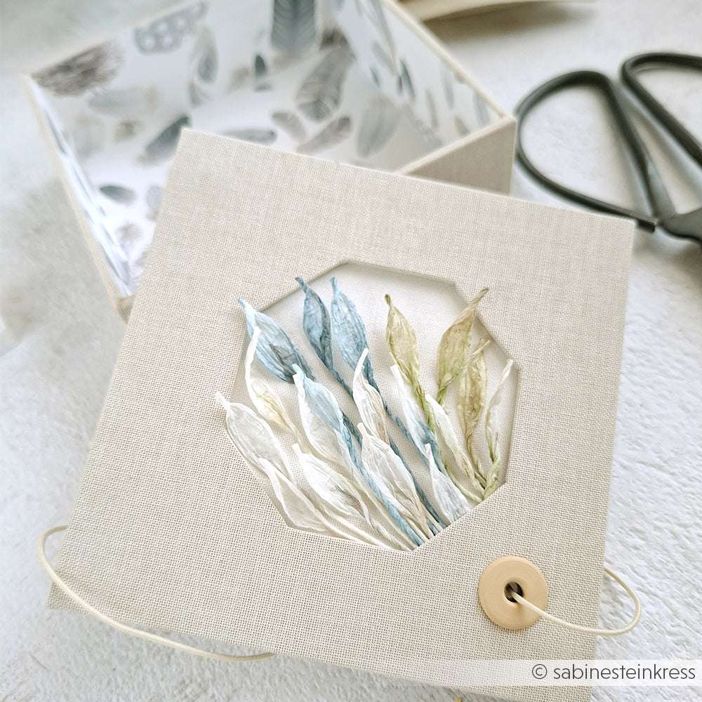 Eine beige Geschenkverpackung von Paperphine mit Knopfverschluss, verziert mit zarten blauen, grünen und weißen Stoffblättern - ideal für Bastelprojekte oder besondere Geschenke.