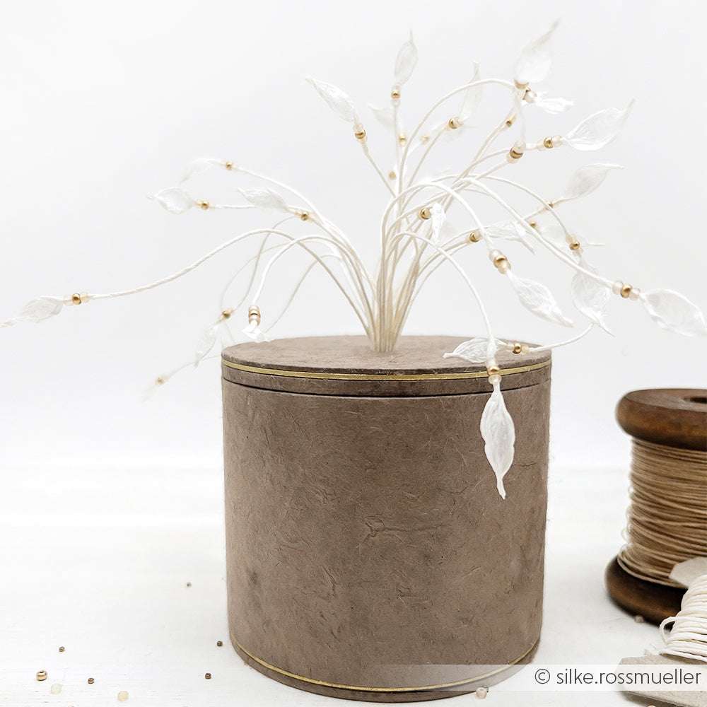 Zarte Paperphine-Zierzweige mit durchscheinenden Blättern in einer runden braunen Schachtel; daneben eine Spule Paperphine-Papierschnur - ideal für Bastelprojekte oder zum Verpacken von Geschenken.