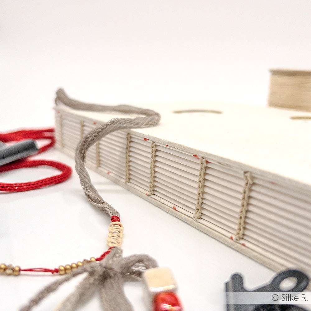 Ein handgefertigtes Sajou-Buch mit freiliegender Rückennaht, hergestellt aus gewachstem Leinen und Fadenbindungen, begleitet von dekorativen Perlenschnüren.