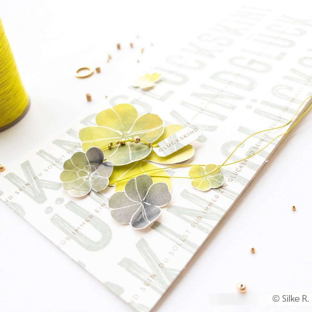 Eine Karte mit Papierkleeblättern, Goldperlen und deutschem Text ist mit Sajou gewachstem Leinen und gelbem Garn verziert, daneben liegt eine Spule Sajou Leinengarn.