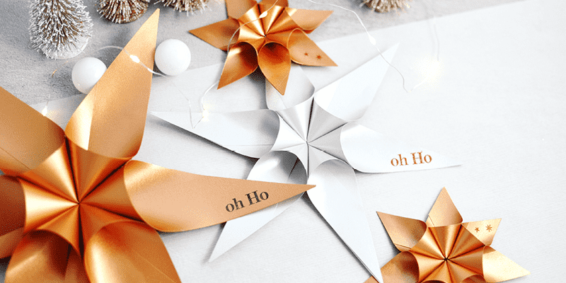 Gold- und silberfarbene Papiersterne mit "Oh Ho"-Text, umgeben von festlichen Dekorationen auf weißem Untergrund.