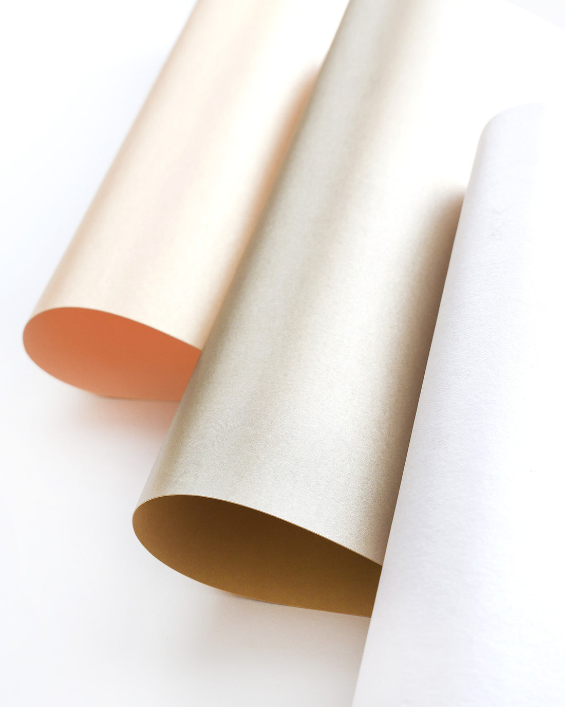 Drei Papierbögen in Beige, Gold und Weiß, leicht gerollt und auf einer weißen Fläche angeordnet.